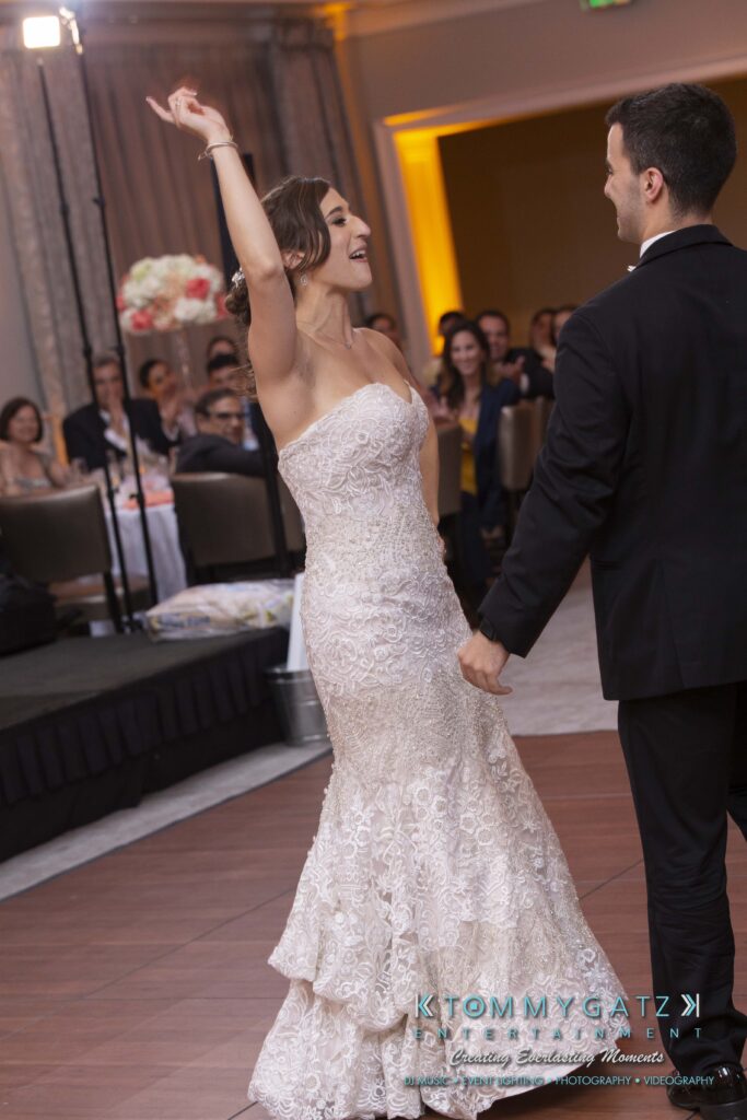 Bride and groom dance joyously at their wedding reception, surrounded by seated guests in an elegantly decorated venue.