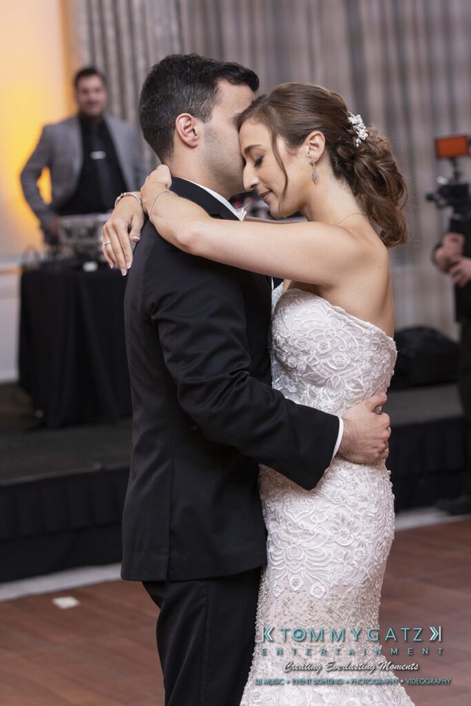 A bride and groom share a dance at their wedding reception. The bride wears a strapless lace gown, and the groom is in a black suit. A DJ stands in the background.