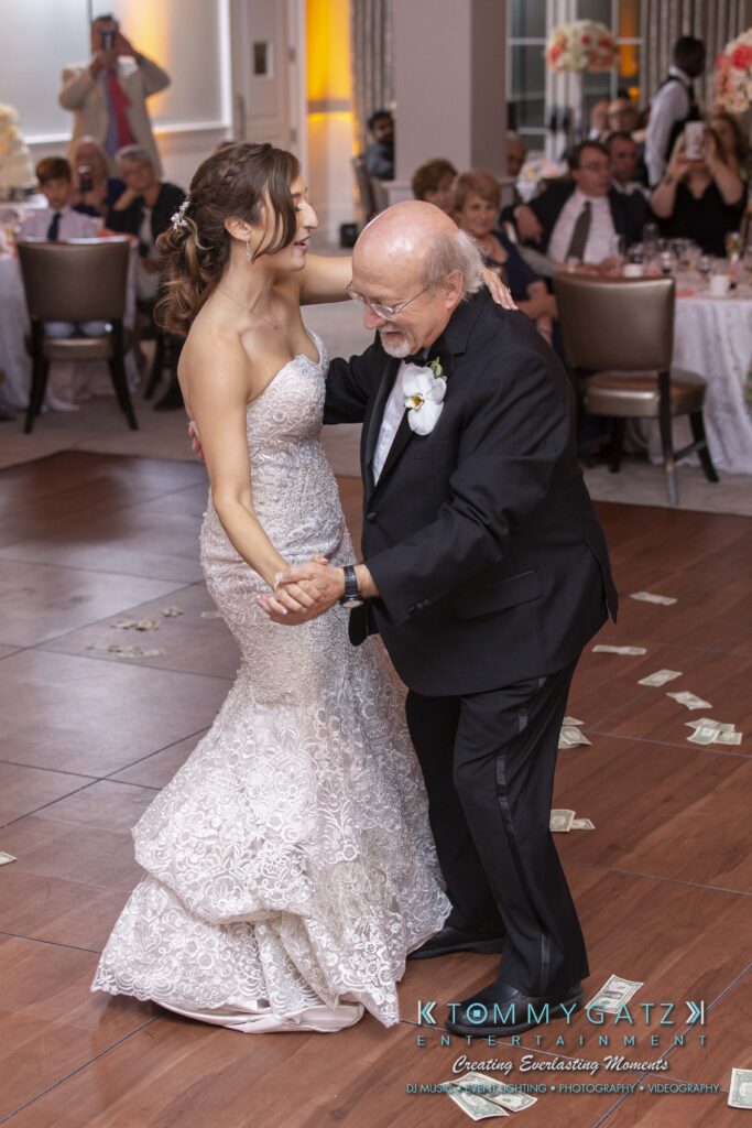 A bride in a lace dress dances with an older man in a suit and bow tie on a wooden floor, surrounded by guests.