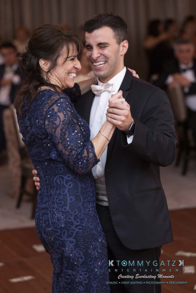 A man and woman, dressed formally, smile and dance together indoors at an event.