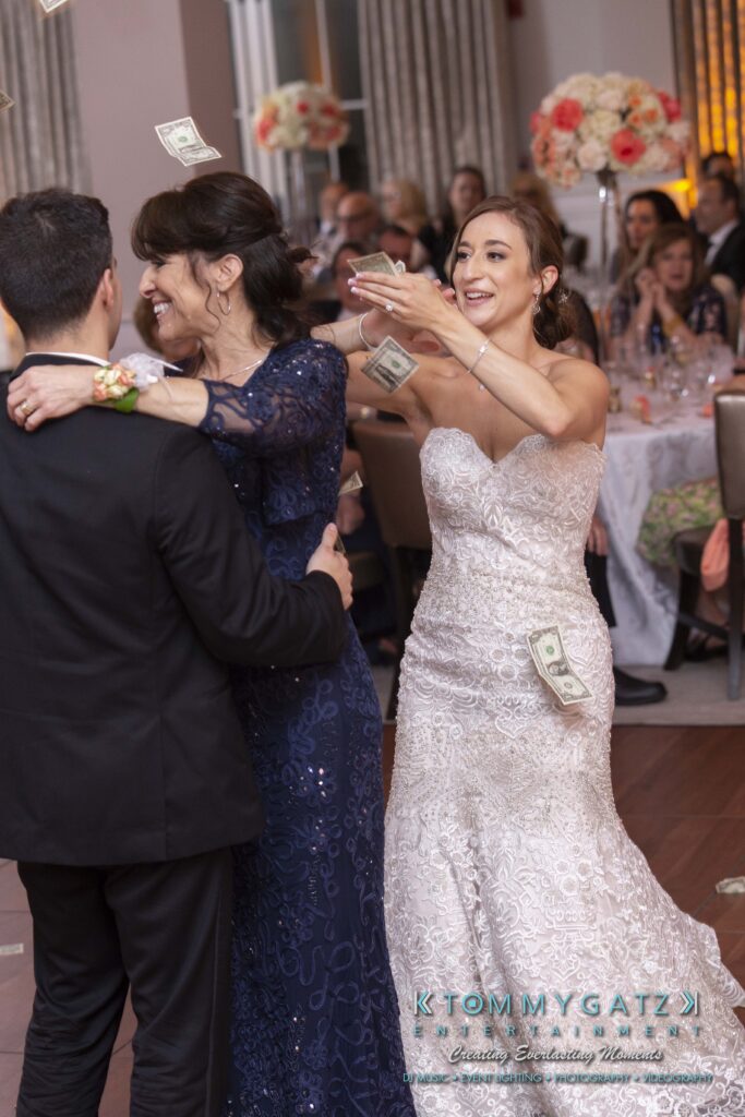 Urania-Aristidis-060218-RECEPTION-Low-Res-WM-901 - TOMMY GATZ ENTERTAINMENT A bride in a white dress places a dollar bill on a man dancing with a woman in a blue dress at a wedding reception. Guests are seated at tables in the background.