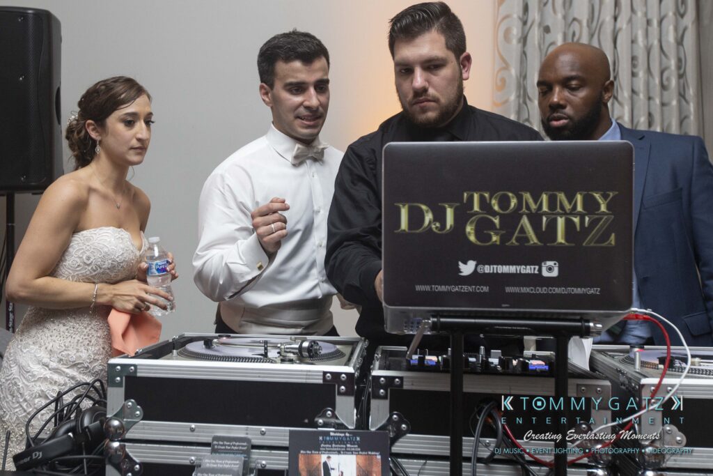 A bride and three men stand around a DJ booth with equipment. One man points while others focus on the laptop screen displaying "Tommy Gatz.