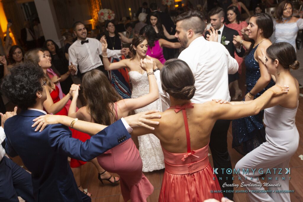 A group of people on a dance floor, holding hands in a circle at an event. They're dressed in formal attire and appear engaged in a lively dance.