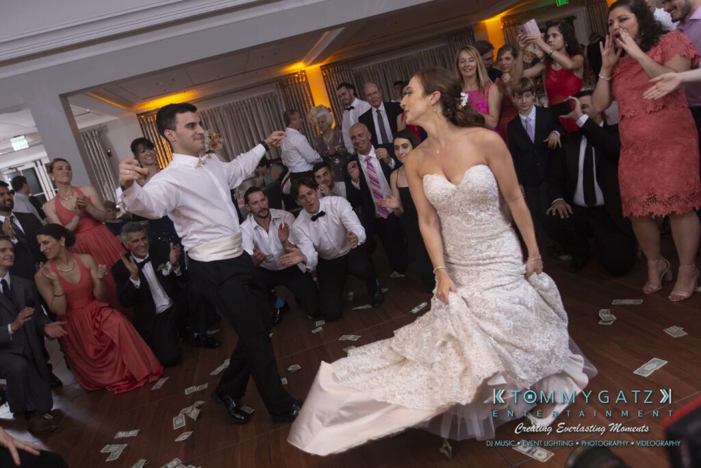 A bride and groom dance at their wedding reception, surrounded by cheering guests. Money is scattered on the floor, and people are clapping and watching with excitement.