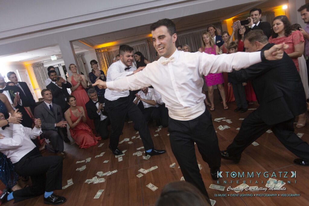 A man in a white shirt and bow tie dances energetically on a wooden floor while guests seated and standing around watch. Dollar bills are scattered on the floor.