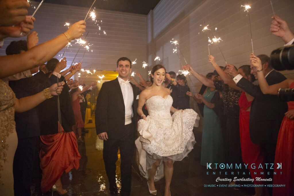 Bride and groom smile while walking through a crowd holding sparklers at an indoor evening event.