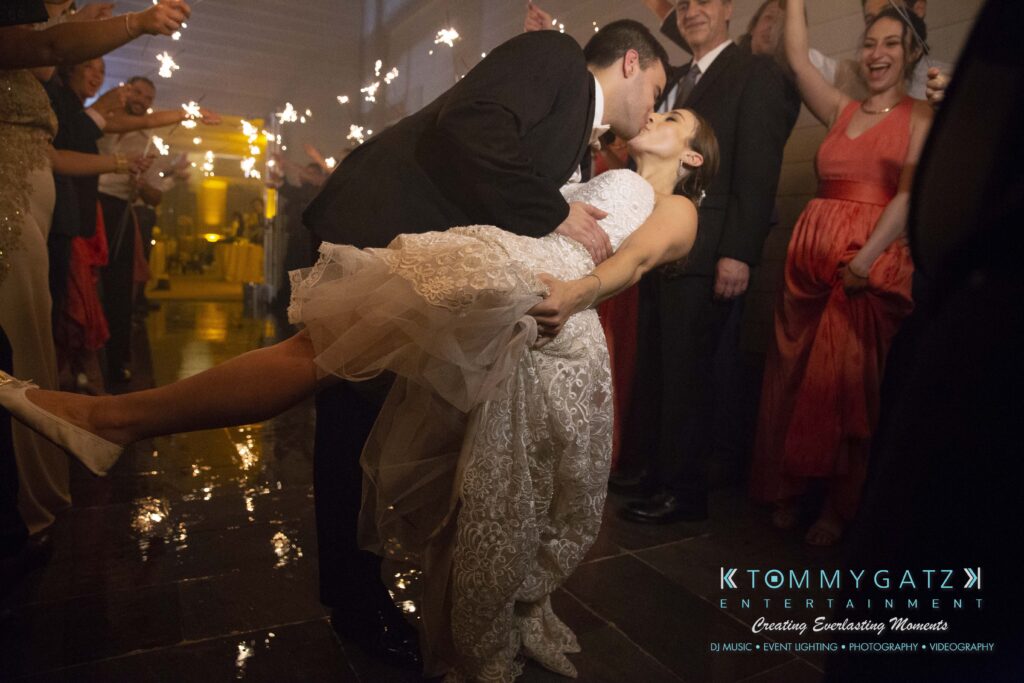 A groom dips the bride for a kiss while guests surround them with sparklers during an indoor celebration.