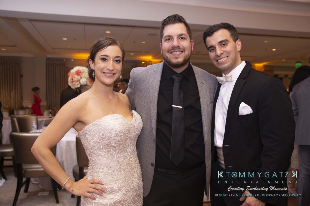 A bride, a man in a gray suit, and another man in a black suit pose together at an indoor event.