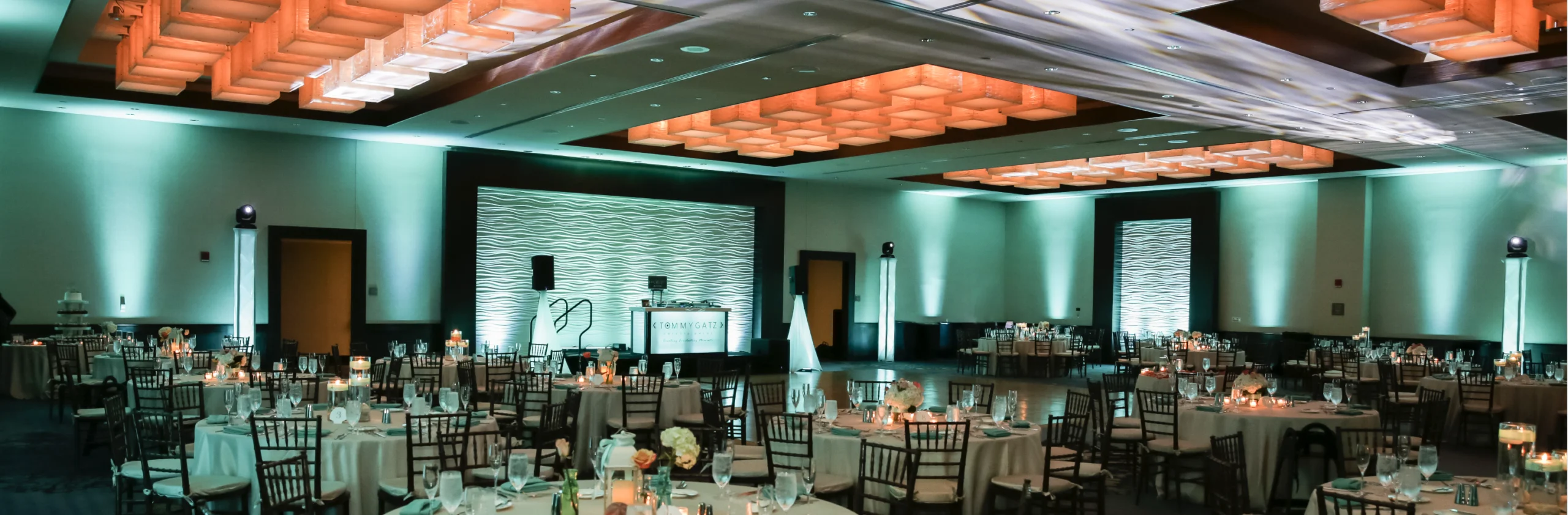 Dimly lit banquet hall with round tables set for an event. Walls and ceiling feature green and orange lighting accents.