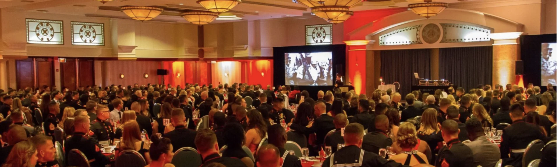 A large group of people in formal attire sit at round tables in a banquet hall with presentation screens and decorative lighting.