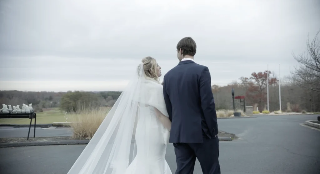 A bride and groom are walking outdoors on a paved path, with the bride in a white dress and veil, and the groom in a dark suit.