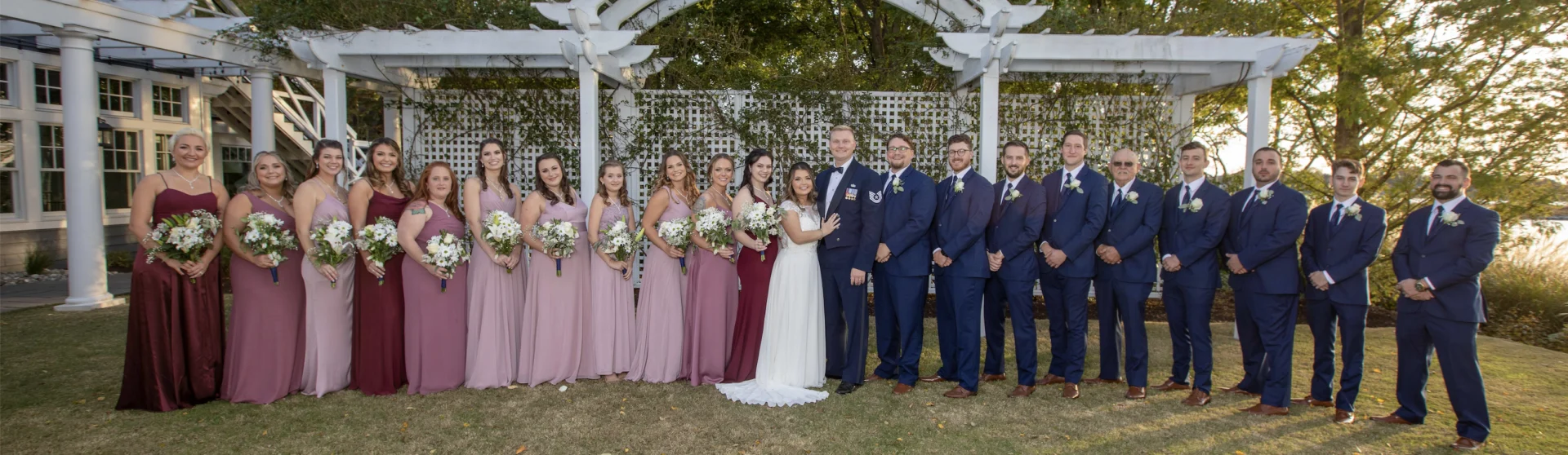 A wedding party poses outdoors under a white pergola. The group includes bridesmaids in pink dresses and groomsmen in navy suits, flanking the couple in the center.