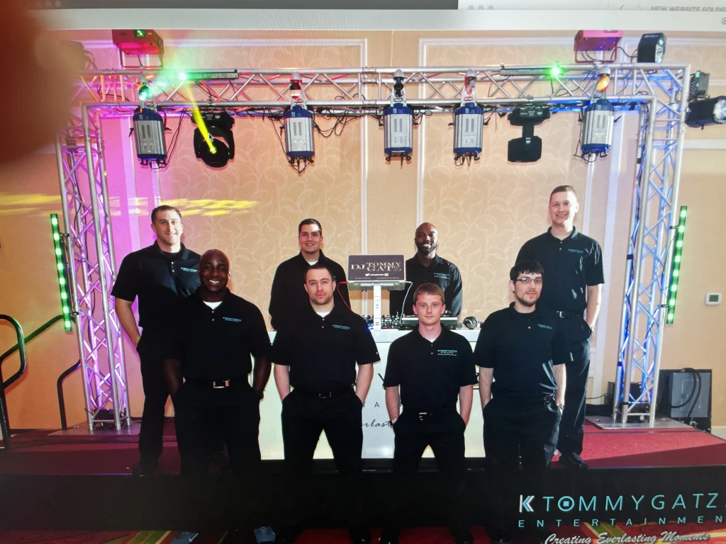 A group of eight people in matching black shirts stands in front of a DJ setup with lighting equipment.