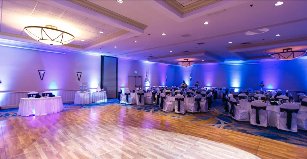 Ballroom set up for an event with round tables and chairs covered in white and black cloth, cake tables on the side, wooden dance floor in the foreground, and purple lighting.