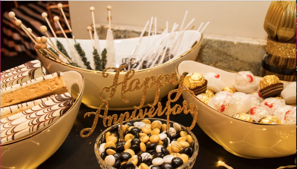 A dessert table with bowls of assorted sweets, including chocolate truffles, cake pops, and candies, features a "Happy Anniversary" sign.