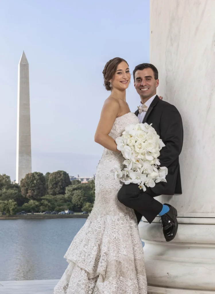 A bride and groom pose by a marble column with the Washington Monument in the background. The bride is holding a bouquet of white flowers.