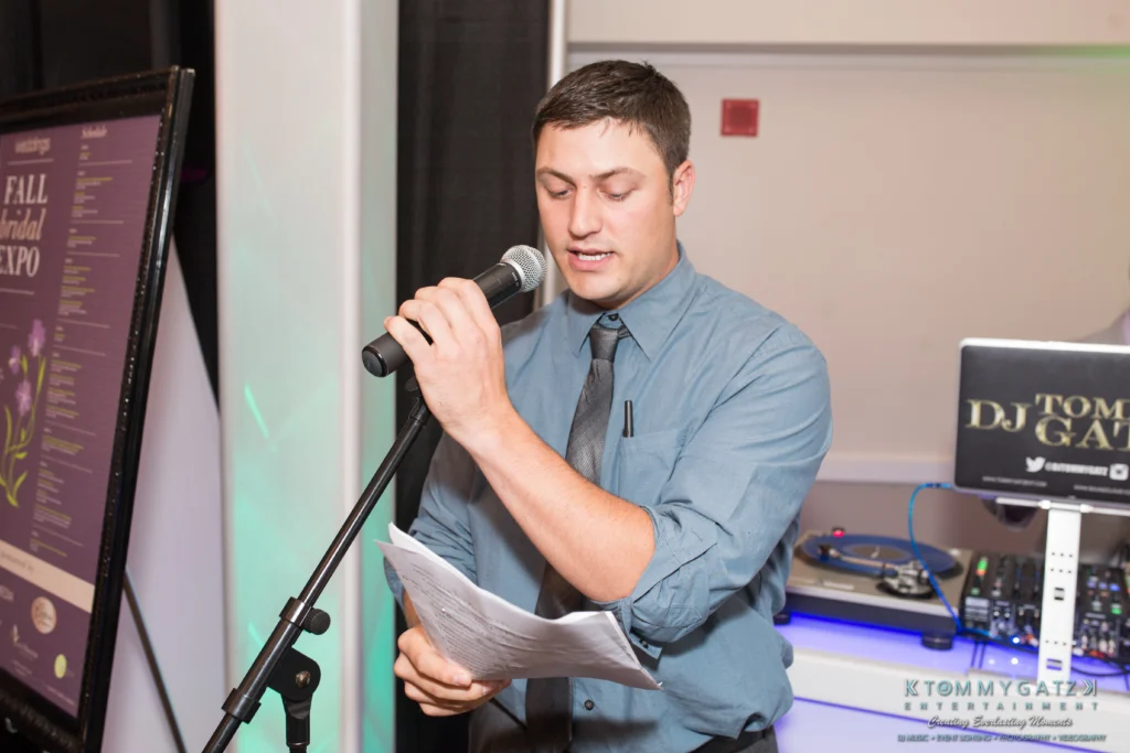 A man in a blue shirt and tie speaks into a microphone while holding papers. DJ equipment is visible in the background.