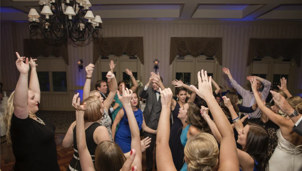 A group of people are dancing enthusiastically with their arms raised in a room with chandeliers and patterned walls.