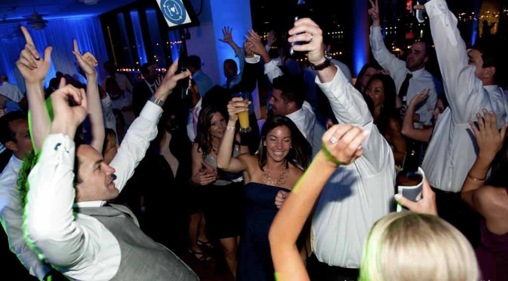 A group of people dancing and raising drinks in a lively party atmosphere with dim lighting and a blue color theme.