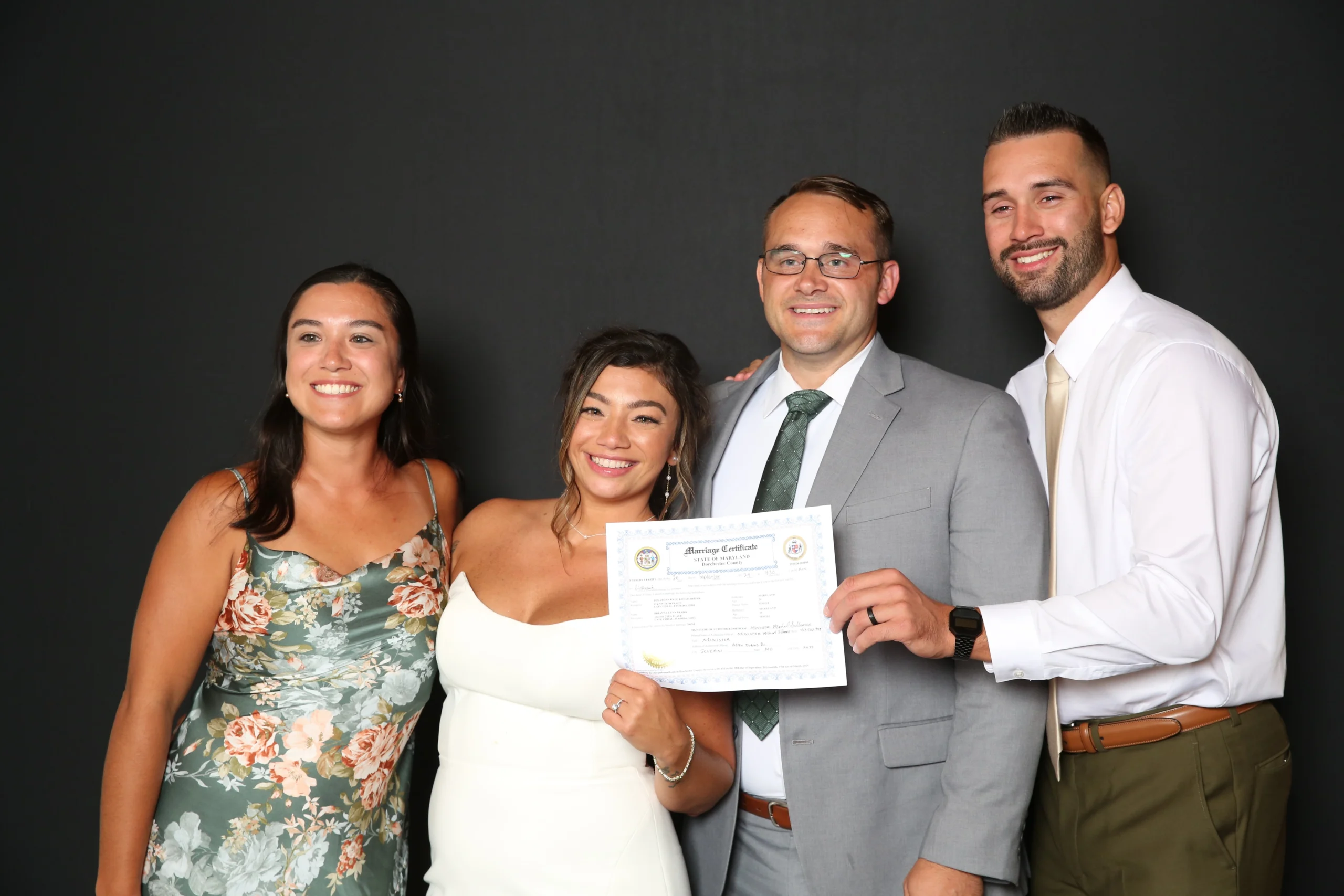Four people pose together; two in dresses and two in suits. One person holds a marriage certificate with a smile. Black background.