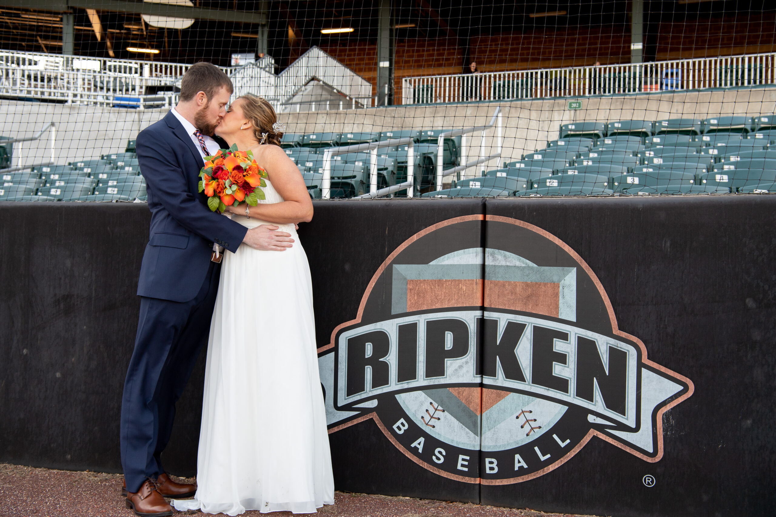 A couple in wedding attire embraces in front of a "Ripken Baseball" logo at a baseball stadium, with the bride holding a bouquet of orange flowers.