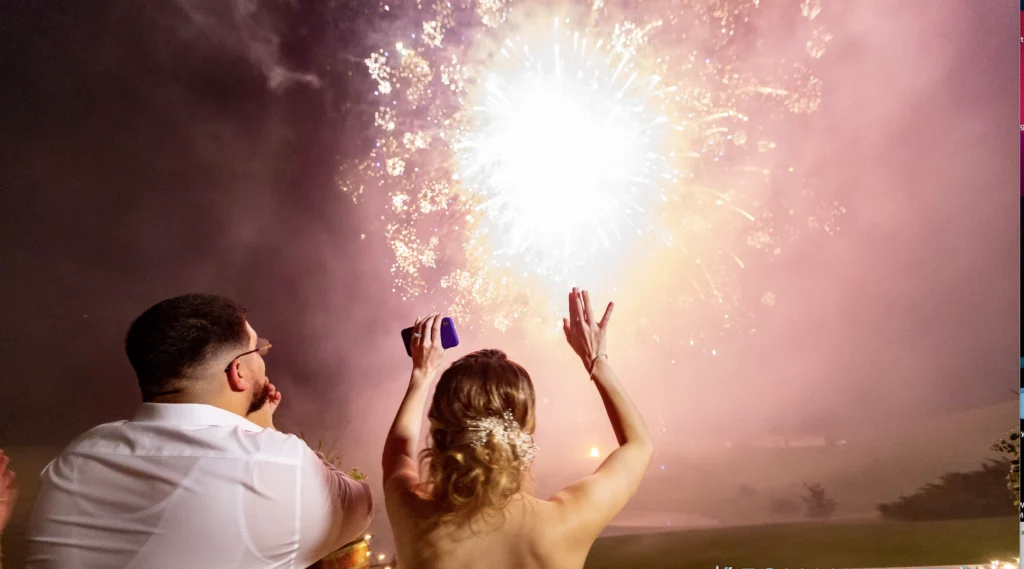 A couple watches colorful fireworks at night, with the woman raising her hands in excitement.