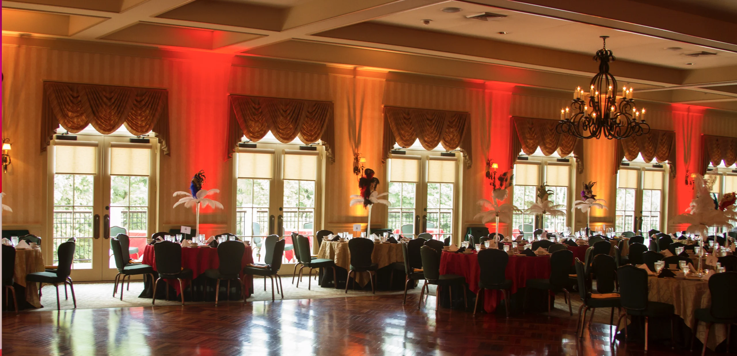 A large, elegant banquet hall with round tables set for dining. The room features red lighting accents, chandeliers, and tall windows with valanced curtains.