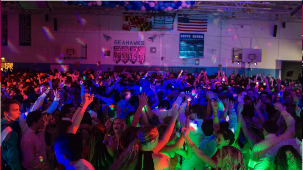 A large group of people dancing under colorful lights in a gymnasium with banners and basketball hoops visible.