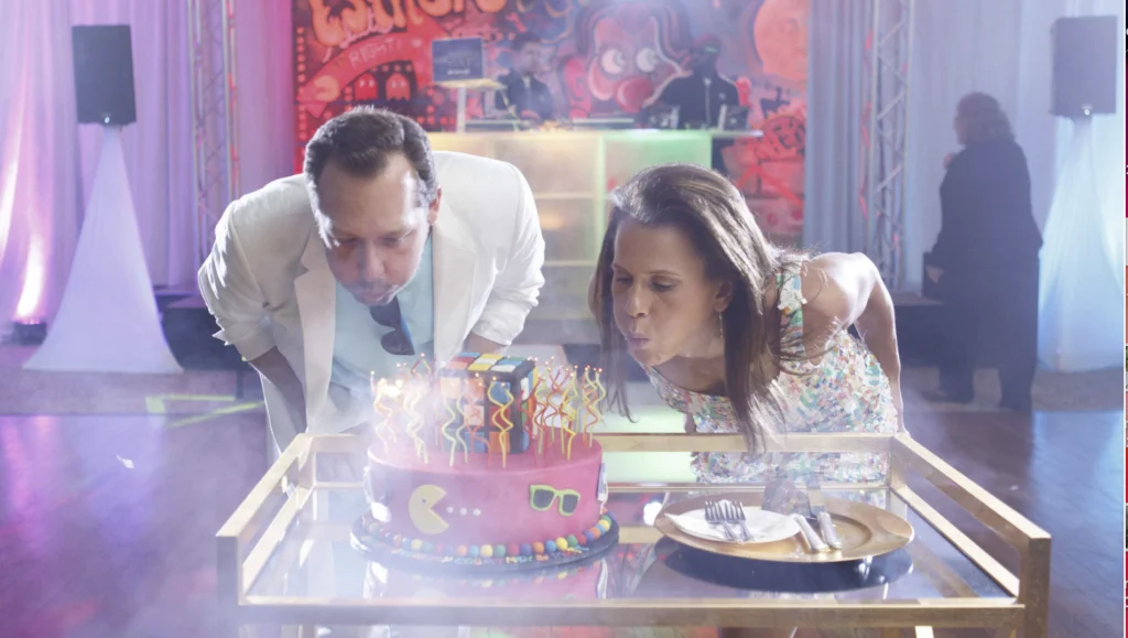 Two people blow out candles on a colorful cake with a Pac-Man design, placed on a glass table. A DJ booth with colorful lighting is visible in the background.