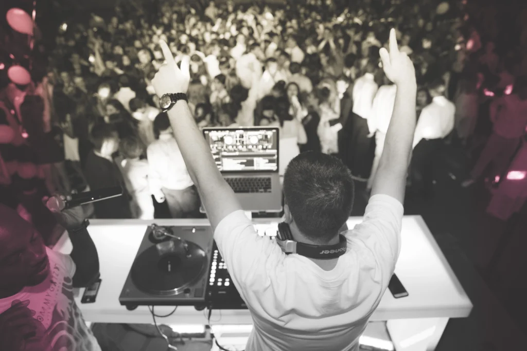 DJ with raised arms performing in front of a large, energetic crowd. DJ equipment and laptop on the table. Scene lit with dynamic lights.