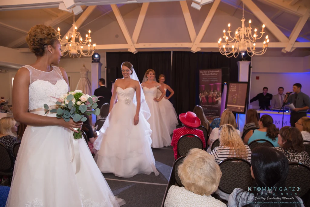 Two models walk on a runway in bridal gowns during a fashion show. Another woman holds a bouquet and observes. An audience watches, seated in front of an elegant room.