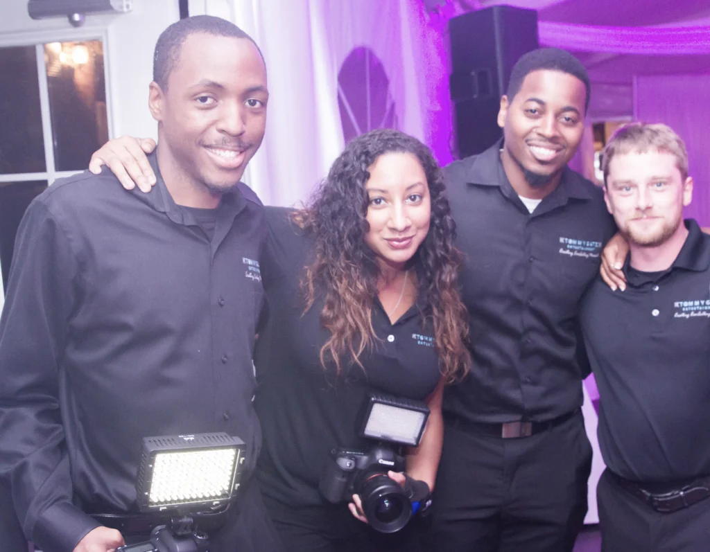 Four people in black attire pose together, two holding cameras with mounted lights, in a well-lit indoor setting.
