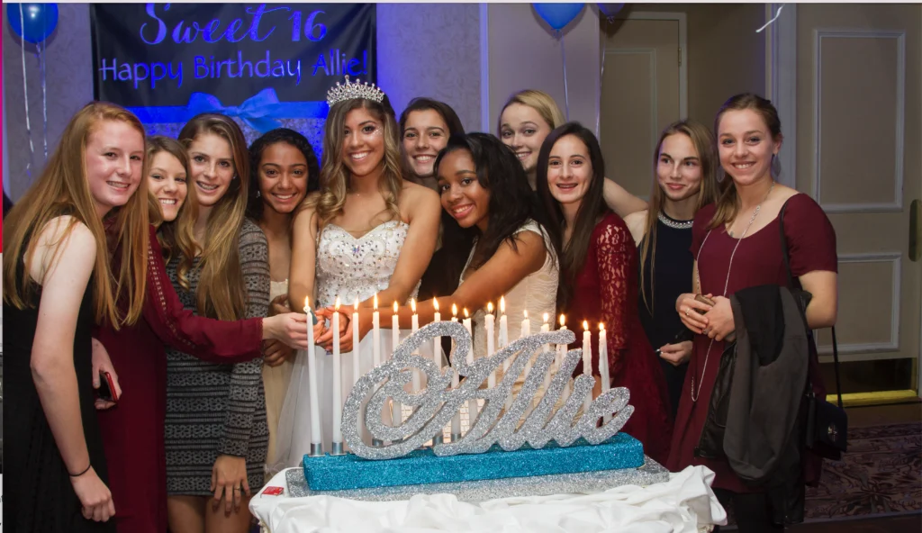 A group of young women pose around a "Sweet 16" cake with lit candles. A banner in the background reads "Happy Birthday Allie!.
