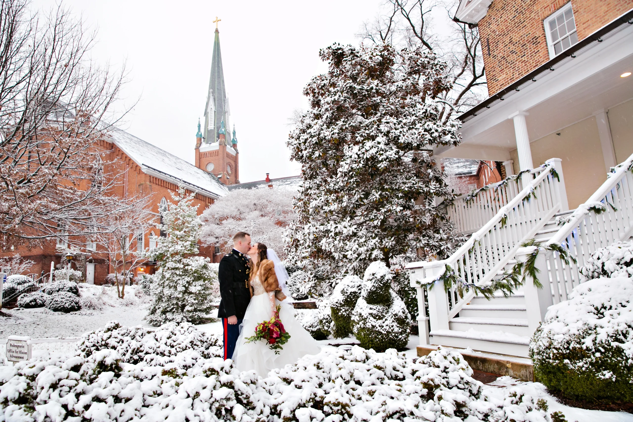 A couple in wedding attire kisses in a snowy garden with a church spire in the background.