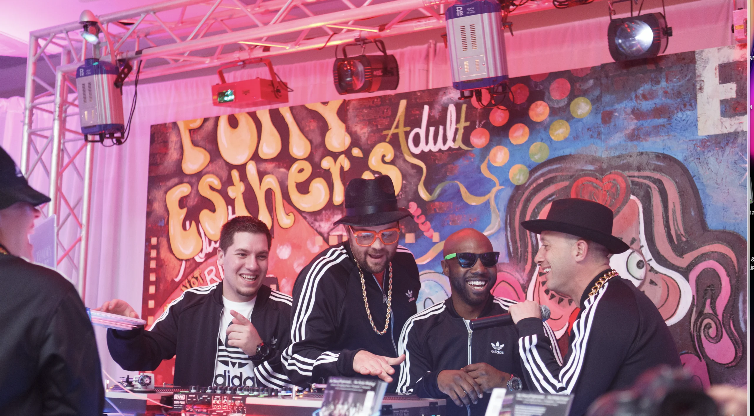 Four men in matching black and white outfits stand at DJ turntables, smiling and talking, with a colorful graffiti wall in the background.