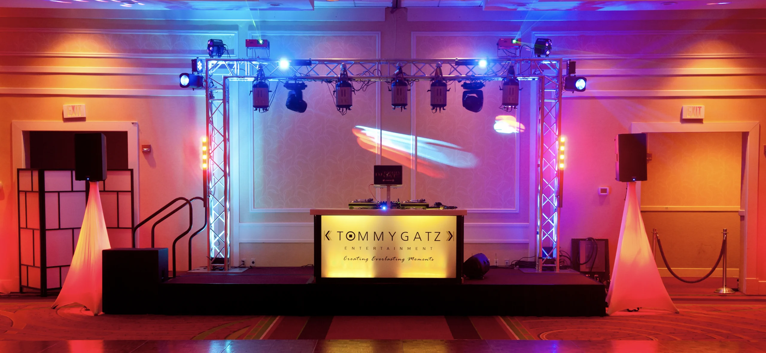 A DJ setup with colorful lights on a stage features two speakers, a truss with spotlights, and a screen projecting moving visuals. A sign reads "Tommy Gatz Entertainment.