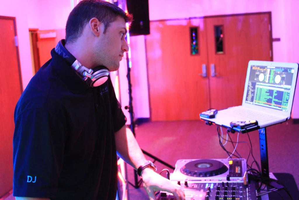 A DJ in a dark room mixes music on a console with a laptop displaying tracks. He wears headphones around his neck, surrounded by equipment and colorful lights.