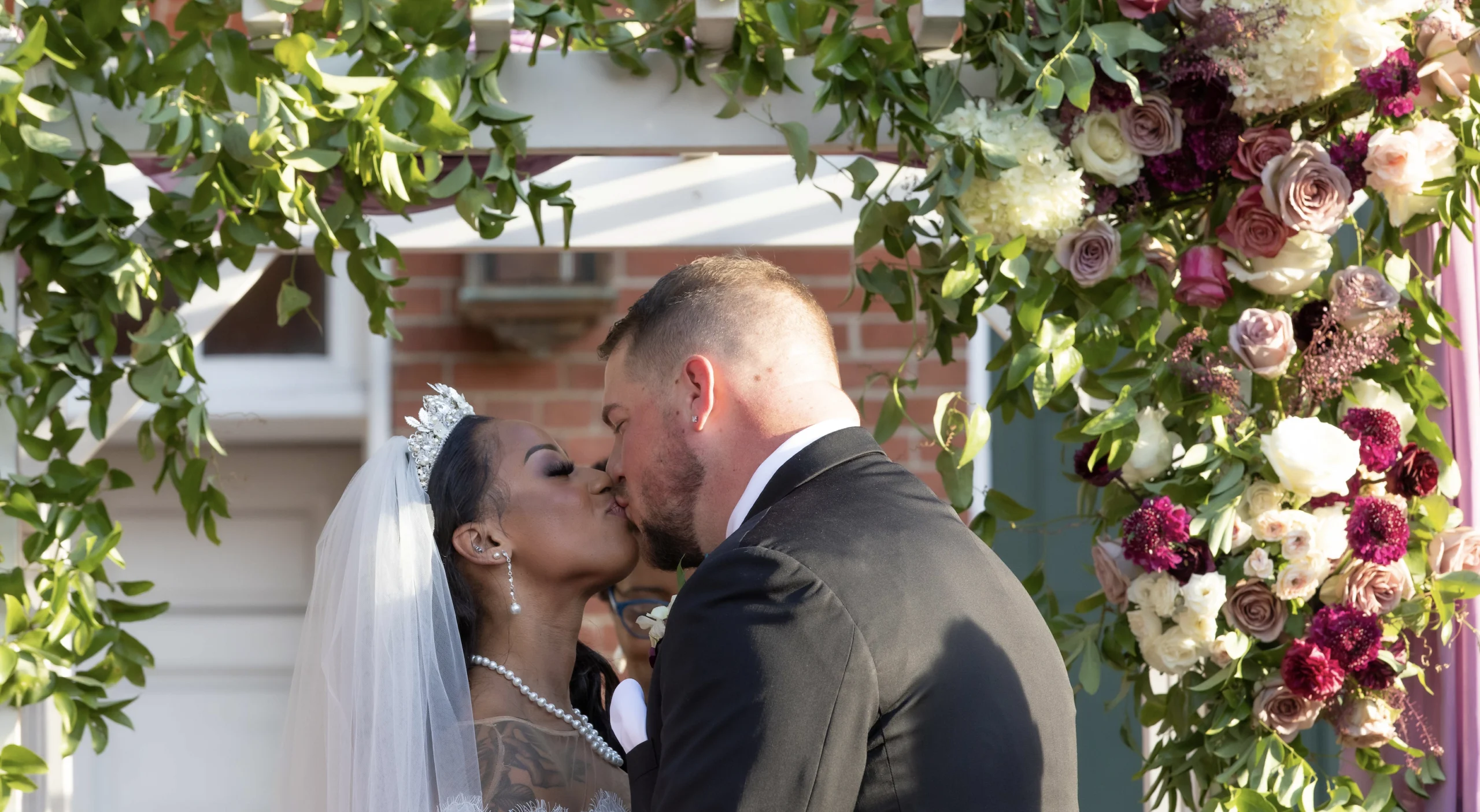 A bride and groom kissing under a flower-decorated archway during an outdoor wedding ceremony.