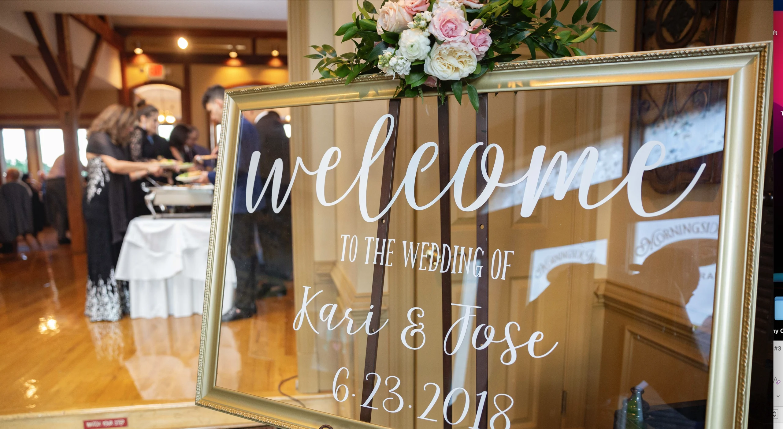 A wedding sign with "Welcome to the Wedding of Kari & Jose 6.23.2018" in white script. In the background, guests are seen at a buffet table.