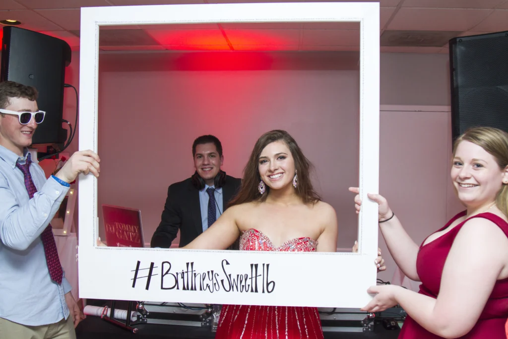 Four young people pose with a large white frame at a party. The frame has a hashtag for "Brittney's Sweet 16." One person in a red dress stands in the center.