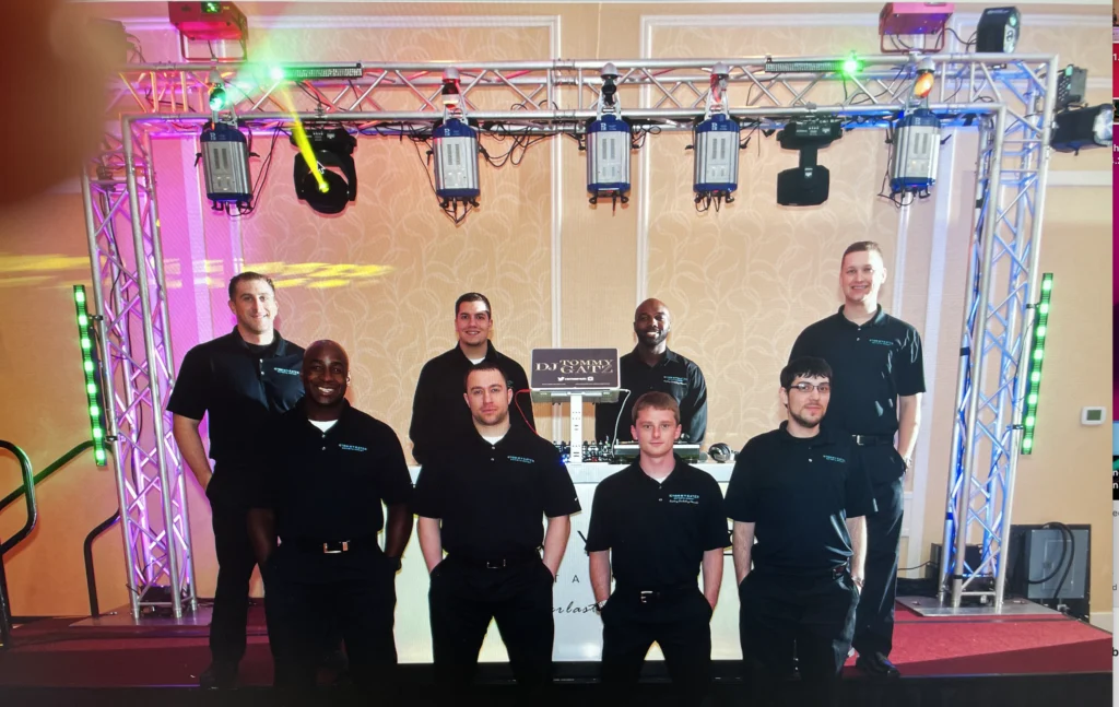 Eight people wearing matching shirts stand in front of a DJ booth and colorful lighting setup.