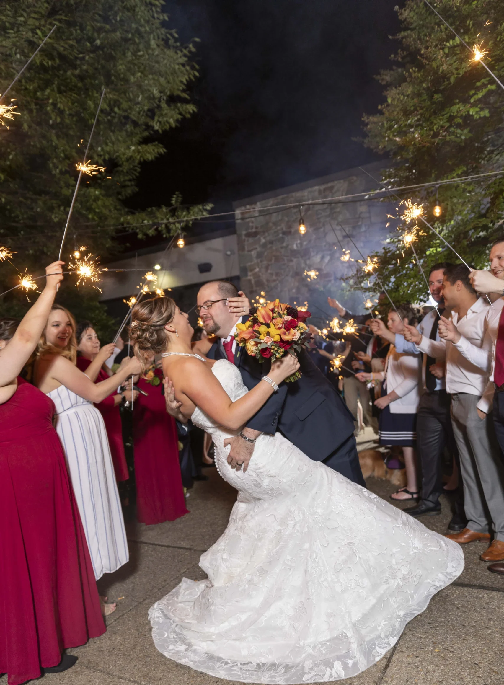 Bride and groom share a kiss surrounded by guests holding sparklers. The bride wears a white dress, and the groom is in a suit. Guests are in formal attire, celebrating outdoors at night.