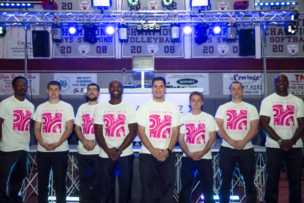 Seven men in matching pink and white shirts pose in front of a brightly lit stage with sports banners in the background.