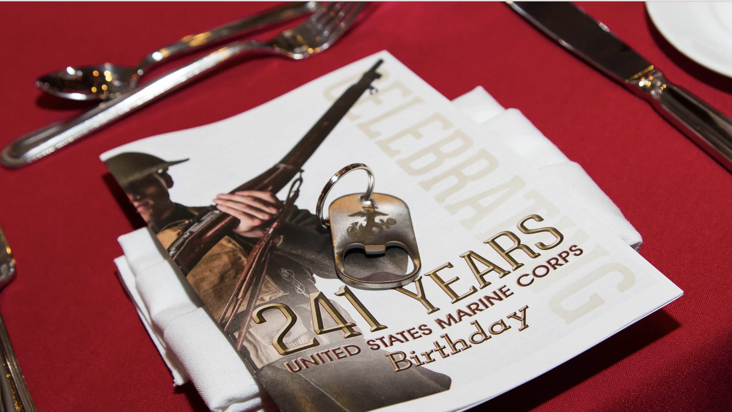 A United States Marine Corps birthday program and dog tag rest on a white napkin, set on a red tablecloth with silverware.
