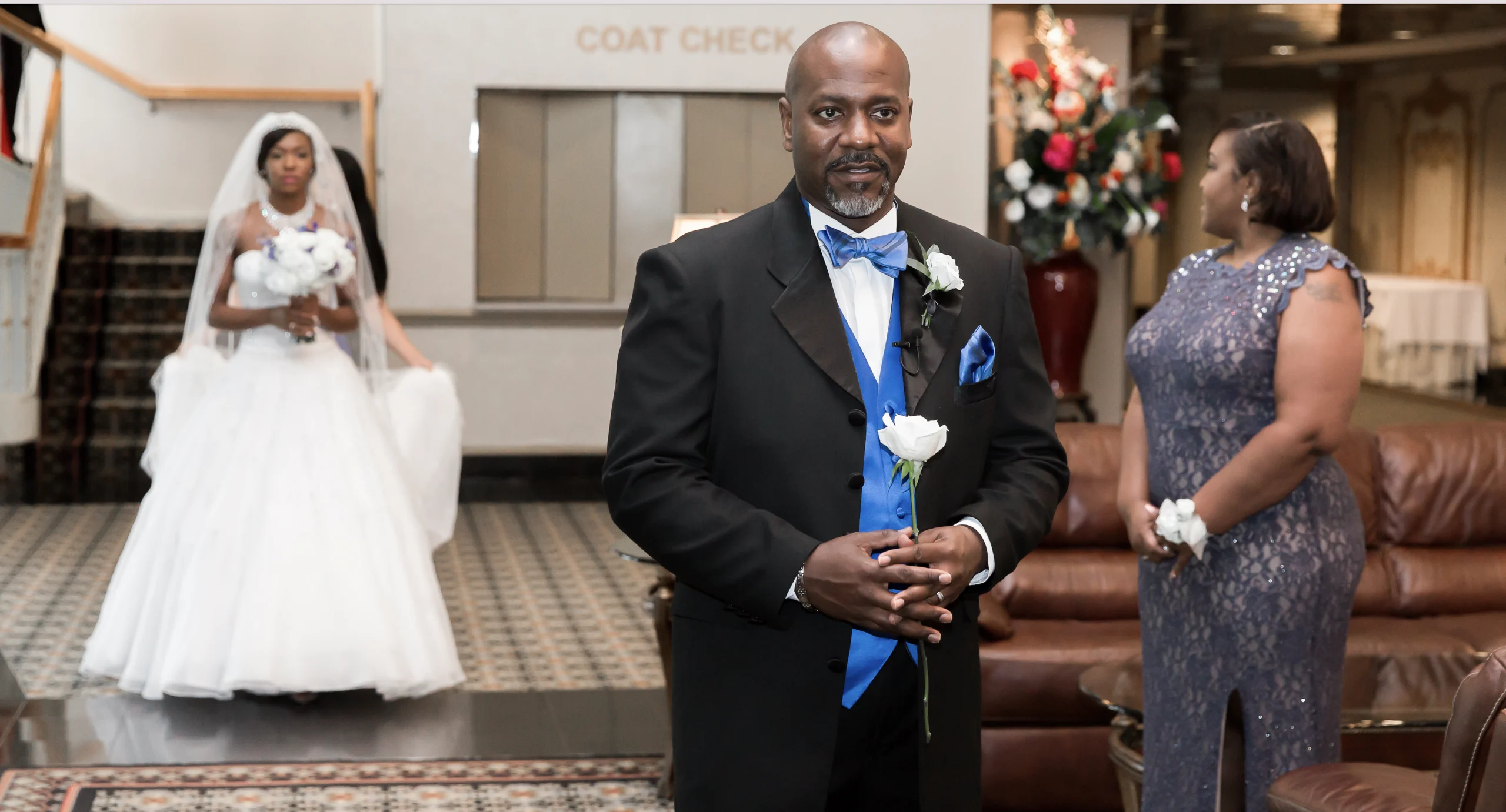 A man in a suit holds a white rose, standing in a lobby. A woman in a gown stands nearby, and a bride walks in the background.
