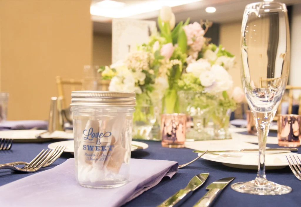 Wedding reception table setting with wine glasses, silverware, a mason jar labeled "Love is Sweet," and a floral centerpiece on a navy tablecloth.
