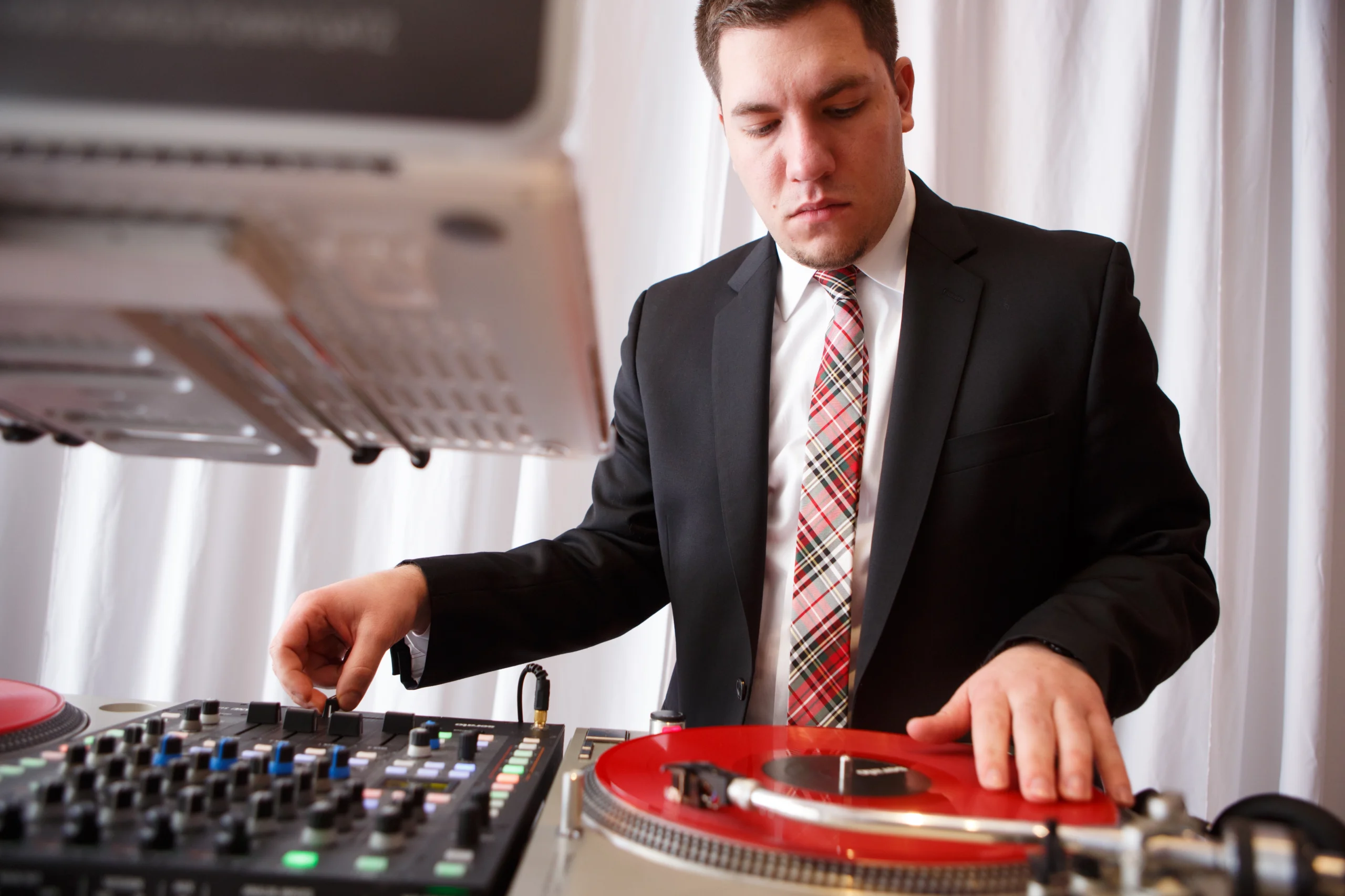 A person in a suit and plaid tie operates DJ equipment, manipulating a red vinyl record and adjusting a mixer.