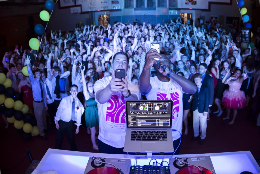 Two DJs take a selfie with a large crowd of excited people at an event. A laptop and DJ equipment are in the foreground.