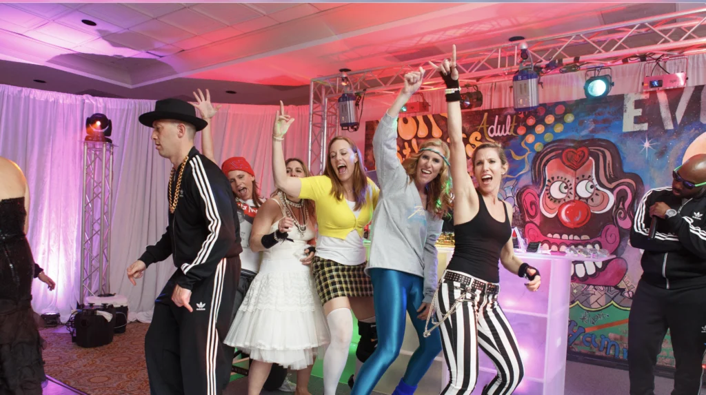 A group of people in colorful outfits and costumes dance energetically on stage. The background features bright lights and a mural of a clown face.