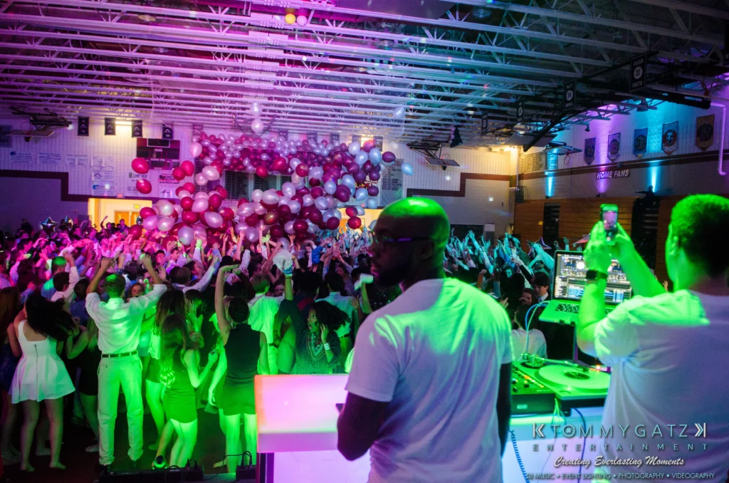 A DJ performing at a lively event in a gymnasium. A crowd on the dance floor is watching balloons drop from the ceiling. Bright stage lights illuminate the scene.